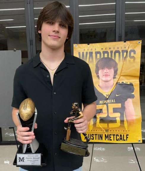 Austin Metcalf in a black shirt, holding two football trophies in front of his Memorial Warriors number 25 banner.