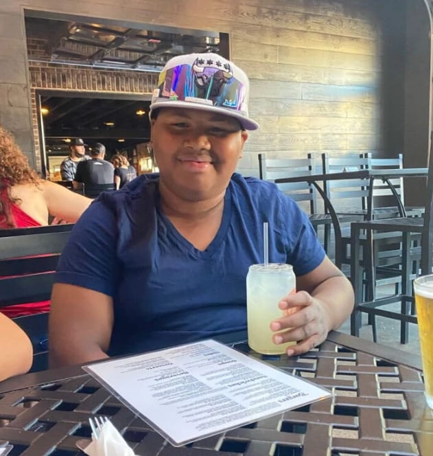 Pierre Johnson, a teenage boy in a blue t-shirt and patterned cap, seated at a restaurant table holding a lemonade.